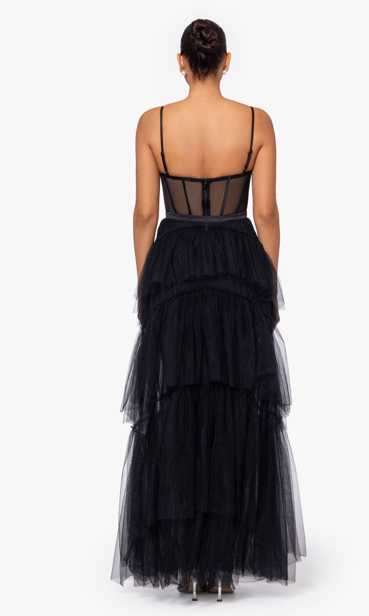 A woman faces away, wearing the Sheer-Waist Long Black Tiered Prom Dress A26249 featuring thin straps, a sheer bodice, and a corset waist that accentuates her silhouette.